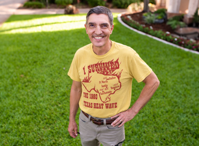 Ken Hyatt, founder of Village Green Lawn and Pest, wearing a vintage 1980 Texas Heat Wave survival shirt, standing in front of a healthy green lawn.