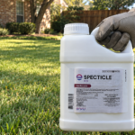 Gloved hand holding a one-gallon jug of Specticle FLO pre-emergent herbicide above a bermuda grass lawn in front of a brick home